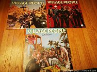 Photo showing three Village People vinyl records with album covers visible, laid out on a wooden floor. Albums displayed are "Cruisin'", "Macho Man", and "Go West".