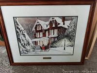 Framed winter scene print depicting a horse-pulled sleigh in front of a large ornate house, snow-covered trees and ground, wooden frame and white mat.