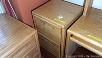 Wooden 2-drawer file cabinet with some wear on top and edges, placed between two other wooden furniture pieces.