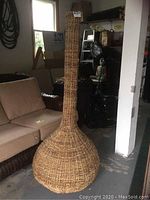 Full view of large tall wicker floor vase with bulbous base and narrow neck, standing upright in room.