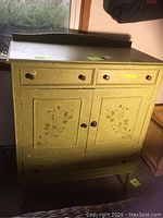 Overall front view of the vintage light green distressed dresser showing two small drawers on top, double doors with floral motifs, and a bottom drawer.
