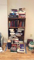 Wide view of wooden bookshelf filled with books including fiction, nonfiction, SAT prep, and cooking titles, plus stacks on the floor.