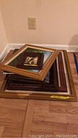 Stack of seven framed wall art paintings on wooden floor, various frame styles and sizes