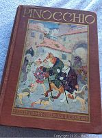 Front cover of antique Pinocchio hardcover illustrated book featuring colorful artwork of Pinocchio and animal characters in a village setting.