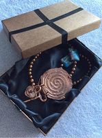 Necklace in gold-tone textured box with black satin lining; shows copper spiral pendant with key and heart charms hanging, necklace beads including copper, jasper, and pearls.