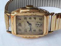 Close-up of rectangular gold-tone Modera wristwatch face, showing worn dial with black Arabic numerals, blued hands, and seconds subdial at 6 o'clock. Watch case has visible surface wear and patina.