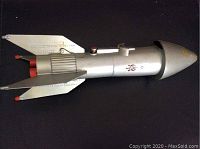 Full side view of the vintage steel space ship coin bank showing silver metal body, fins, and red details.