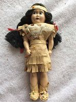 Front view of the full doll showing the tan suede dress with fringe and beadwork, black yarn hair in pigtails, and moccasin boots with bead decor.
