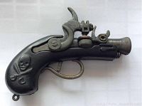 Left side view of the miniature metal pirate cap gun showing skull and crossbones emblem on black grip and metallic barrel and hammer.