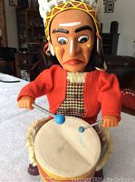 Front view of Native American drumming toy showing facial features, headdress, red jacket, and drum with painted design.