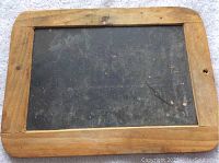 Antique student chalkboard showing one side of the black slate writing surface and wooden frame with rounded corners and signs of wear.