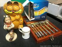 Photo of entire lot arranged on a surface including Garfield figurine, boy figurine, mini cup, Intel processor box, and drill bit set in wooden box.