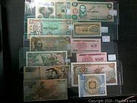 Photo showing 12 world banknotes from different countries including US, Jamaica, North Korea, Japan, Kyrgyzstan. Various colors and denominations visible. Notes are in plastic protectors.