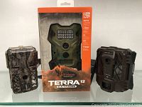 Three Wildgame Innovations Terra 12 Extreme trail cameras, one in original retail packaging, two out of packaging, camouflage design.