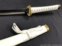 Close-up of white handle with textured wrap, gold guard, and white sheath with braided cord and gold end cap.