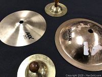 Clear top-down view of an AAX cymbal and a Sabian cymbal with two small brass cymbals, all with wooden handles, showing shiny surfaces and central mounting holes.