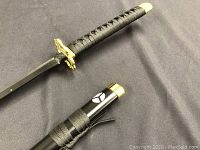 Black sword and sheath laid on black fabric, showing handle wrapped in black cord, gold tsuba, and sheath with white symbols and gold caps.