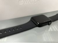 Black Apple iWatch front face with black sport band, displayed on white surface with Apple Watch box in background.
