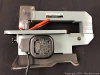 Side view of the Delta 13-inch scroll saw showing metal frame, black base with ventilation, orange blade guard, and clear plastic safety shield.