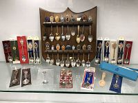 Full lot view showing wooden display plaque with 16 mounted spoons and additional boxed souvenir spoons on glass shelf below.