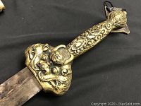 Close-up of sword handle showing detailed ornamental designs in yellow metal with a face motif on the guard.