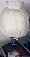 Full view of vintage table lamp showing cream-colored ruched fabric shade with fringe and decorative ceramic base on wooden side table.