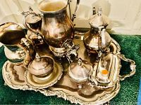 Eight silver plated serving pieces on ornate tray on green carpet