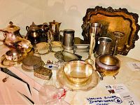 Full view of the assorted silverware including teapots, trays, creamers, plates, and other serving items arranged on a kitchen counter.