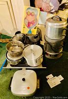 Photo showing assorted cookware including pots, colander, steamer on black stand, electric grill, and bikini cake pan mold in packaging