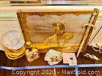 Photo showing a Western-themed plaque with cowboy on horseback, coiled rope, decorative bottle, painted rock, and metal horse lighter arranged on a table.