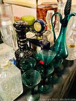 Photo showing a collection of various barware including multiple decanters of different colors and designs, small green glassware, and a tall green glass pitcher with a curved handle.