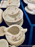 Photo shows stacks of plates, saucers with a delicate pink floral pattern, a cream pitcher, some teacups and saucers arranged on a blue cloth background.