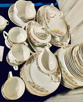 Photo showing multiple pieces of white porcelain china including cups, plates, creamers, and a tureen, all decorated with floral motifs and gold trim.