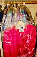 Photo showing women's clothing on hangers covered in dry-cleaning plastic bags, with assorted colors and fabrics, including bright pink and patterned pieces.