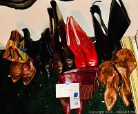 Seven pairs of women's boots arranged on green carpet, showing different styles and colors including red, black, brown, tan, maroon, and multi-colored.