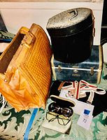 Photo showing two large vintage travel bags, a black vanity case, a white striped belt, and two pairs of glasses along with soft and hard cases on patterned carpet.