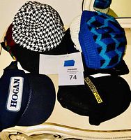 Top view of five casual hats arranged on a surface including a houndstooth patterned cap, blue and black knit cap, white knit cap, black cap with 'DEAL 1 ON 1' patch, and navy baseball cap with 'HOGAN' logo patch.