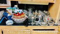 Wide shot showing multiple glass and kitchen items on wooden shelf including glass jars, bowls, an electric appliance, and serving dishes.