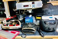 Photo of various electronic and household items on a black surface, showing a black clock radio with CD player, a vintage iron, small clock, handheld electronic device, and accessories.