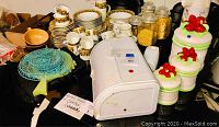 Photo showing an assortment of dishes including patterned cups and plates, a white toaster, green glass plates, ceramic canisters with red fruit decorations, and glass jars with dry goods.