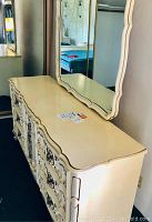 Photo of cream-colored antique style dresser with decorative drawer fronts and matching mirror attached.