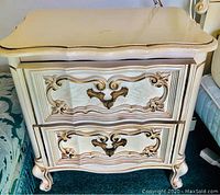 Photo showing an antique style nightstand with cream and gold carved wooden drawers and glass top, ornate drawer handles, and cabriole legs.