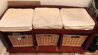 Front view of the wooden bench with all three beige padded seat cushions placed on top and the three wicker baskets underneath, showing wear and soiling on cushions.