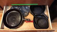 Drawer containing multiple cast iron pans including a large 12.5 inch skillet, a cast iron muffin baking mold, a griddle, and a smaller ridged frying pan with red handle.