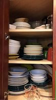 Three shelves inside a cabinet showing ceramic dishes, bowls, cups, and plates stacked in piles, along with some glassware.