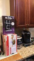 View of four appliances on kitchen counter and boxes. Keurig coffee maker on counter, Koto Plus pressure cooker and EWave convection oven boxed, Mr. Coffee 12-cup coffee maker box visible.