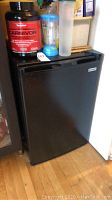 Full view of black Kenmore mini refrigerator with a water bottle and supplement container on top, showing height relative to floor and nearby furniture.