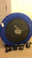 Mini trampoline with blue padded rim and black mat with TÜV NORD GS logo, viewed from front with weights on floor
