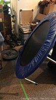 Angled view showing blue mini trampoline with legs, steel springs, assorted weight plates, and dumbbells behind
