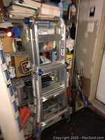 Closed multi-position aluminum ladder standing indoors next to shelves with various items.